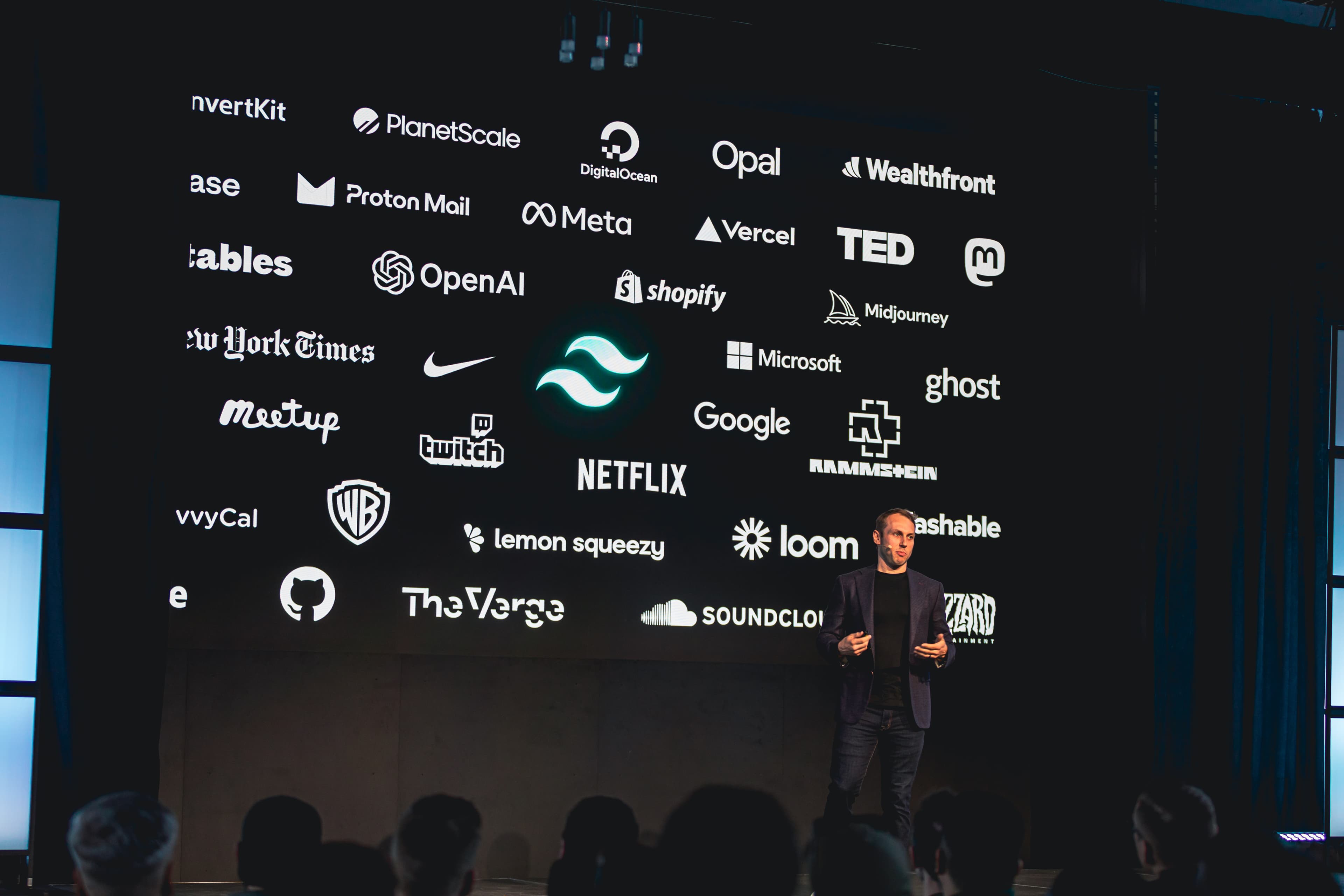 Adam Wathan standing on stage in front of a slide highlighting some of the companies using Tailwind CSS